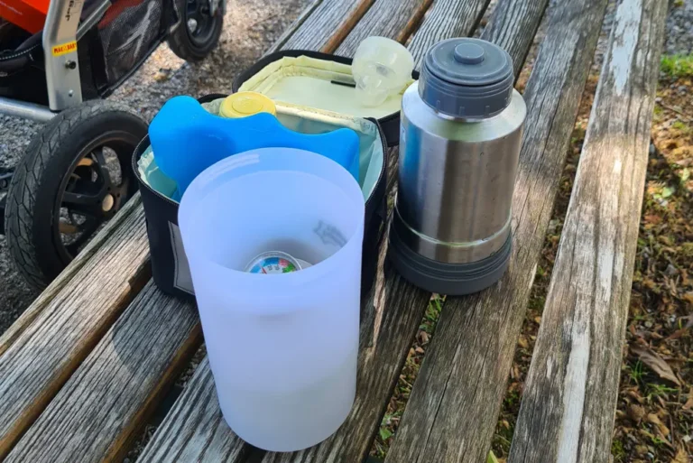 Flaschenwärmer mit Akku und Babyflasche auf einer Parkbank, im Hintergrund ein Kinderwagen