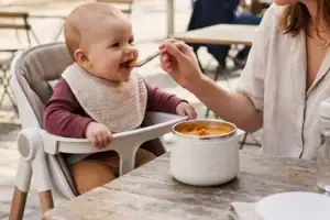 Mutter füttert ihr Baby im Hochstuhl auf einer Café-Terrasse mit Brei aus dem Breiwärmer